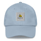 A light blue dad hat with a sun and beach logo embroidered in light blue and yellow on the front. The hat is made of light-colored cotton fabric and has a curved brim. It has an adjustable strap at the back for a comfortable fit."